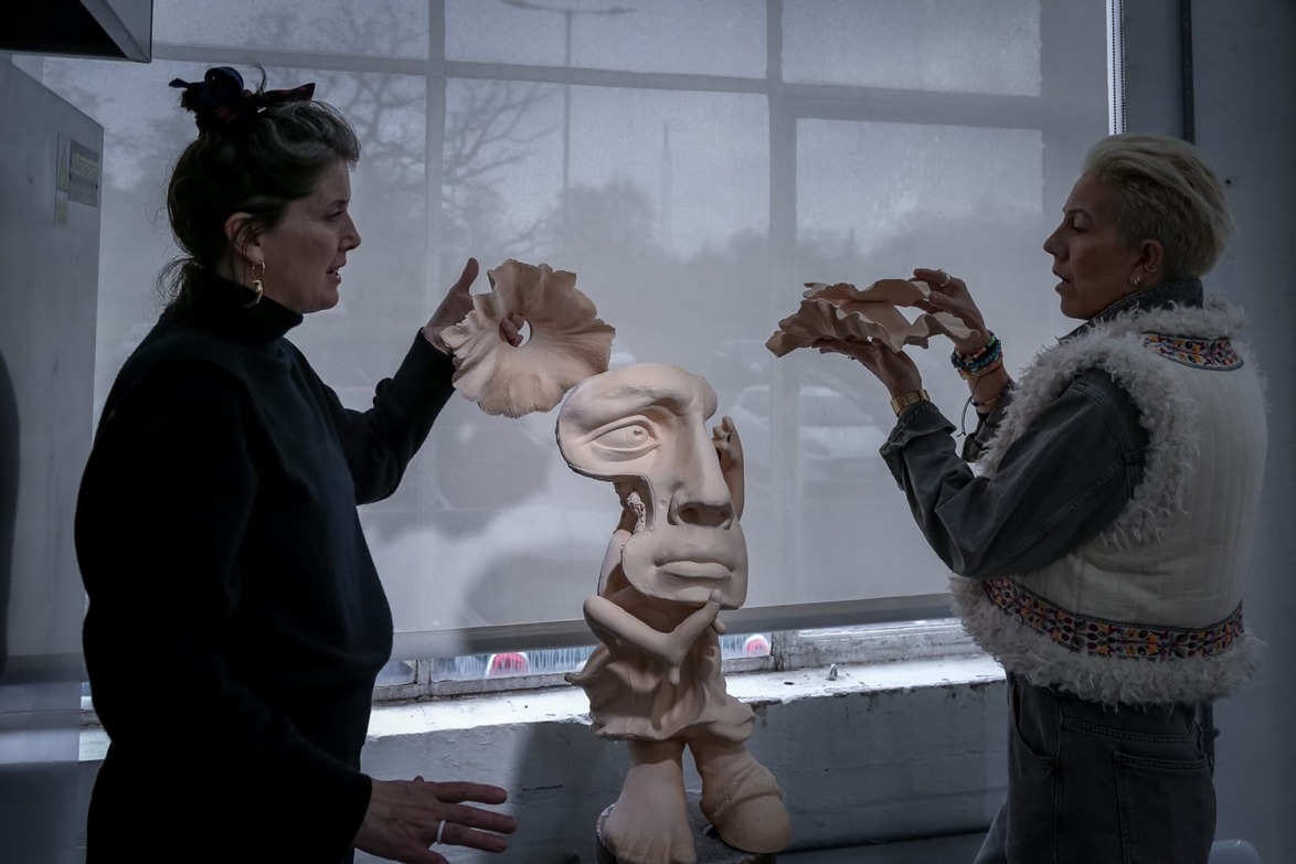 Two people discussing a surreal sculpture with a distorted face in an art studio setting.