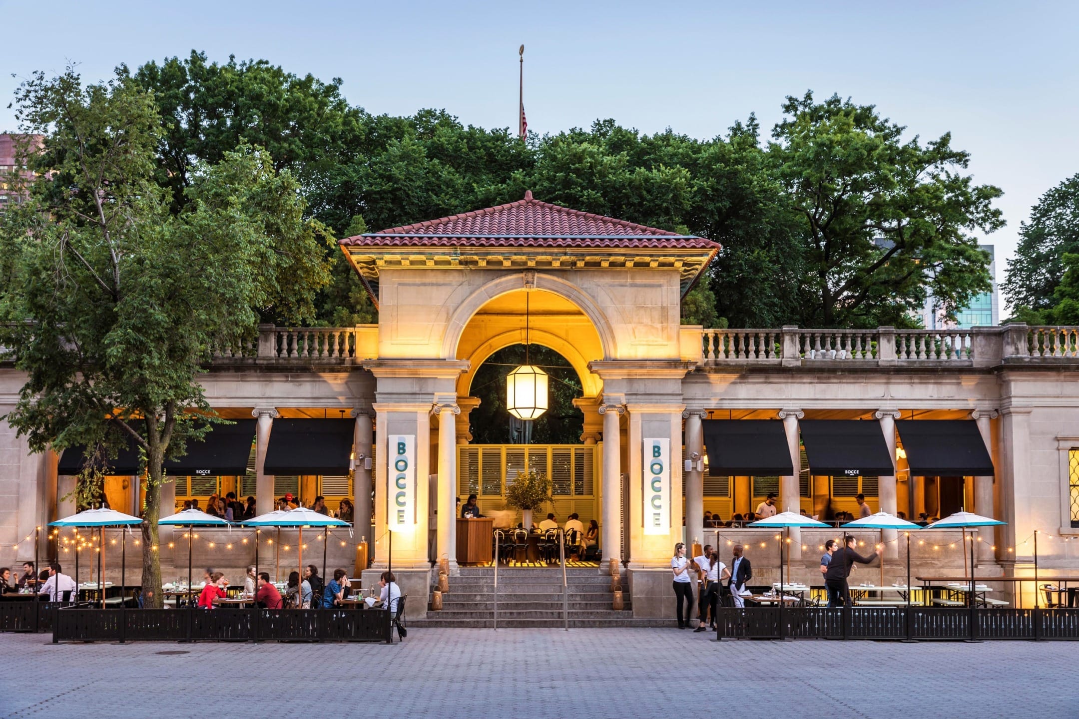 Outdoor dining at an illuminated restaurant with a historic facade surrounded by trees, bustling with people in the evening.