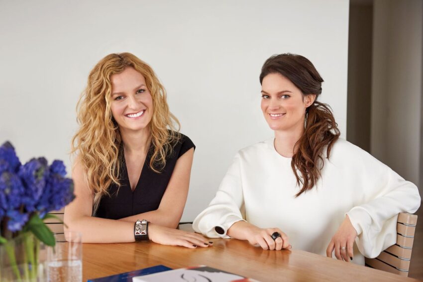 Two women sitting at a table with a bouquet of purple flowers in the foreground.
