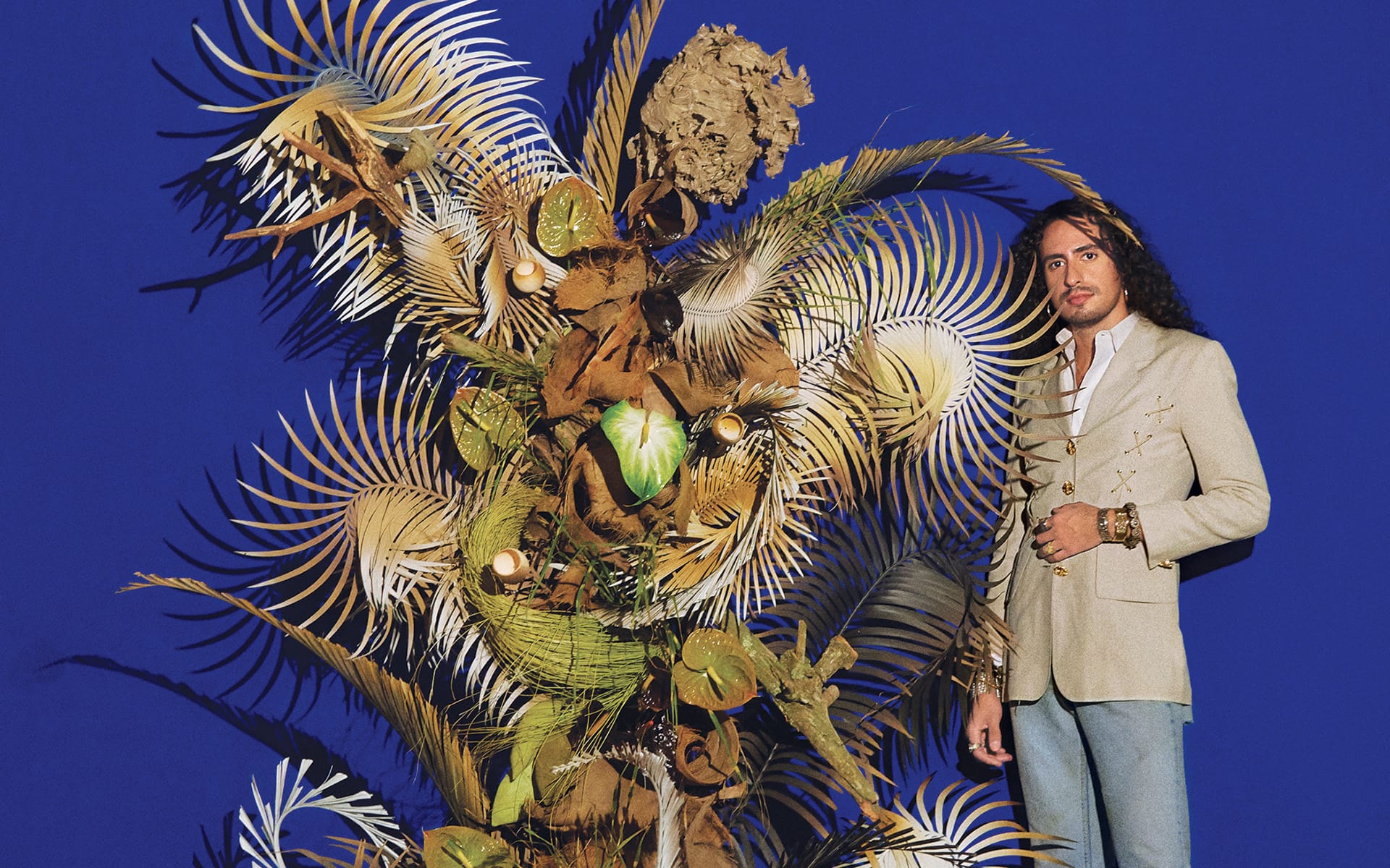 Man in a beige jacket stands beside an abstract sculpture with leaves against a blue background.
