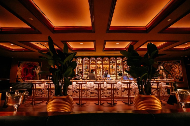 Dimly lit bar interior with cozy seating, decorative ceiling, and illuminated shelves showcasing a variety of bottles.