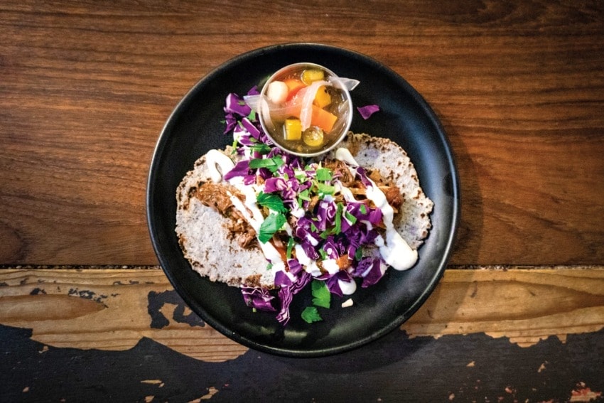 Plate of tacos with shredded meat, purple cabbage, and sauce, served with a side of pickled vegetables on a wooden table.