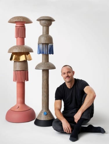Man seated next to two tall, artistic sculptures with intricate patterns and unique textures in a bright studio setting.