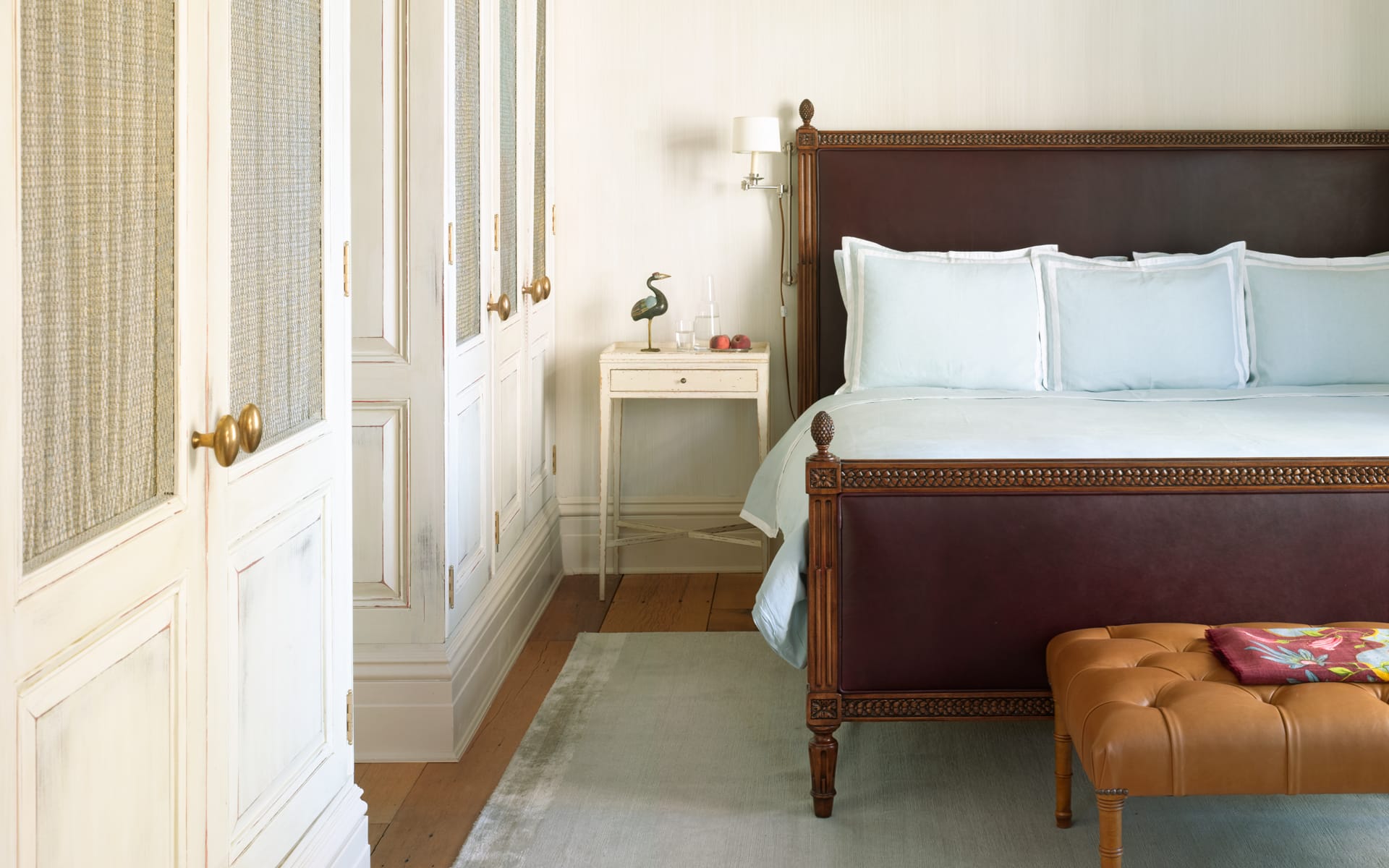 Elegant bedroom with a classic wooden bed, white linens, and a leather bench, adjacent to built-in closets and a bedside table.