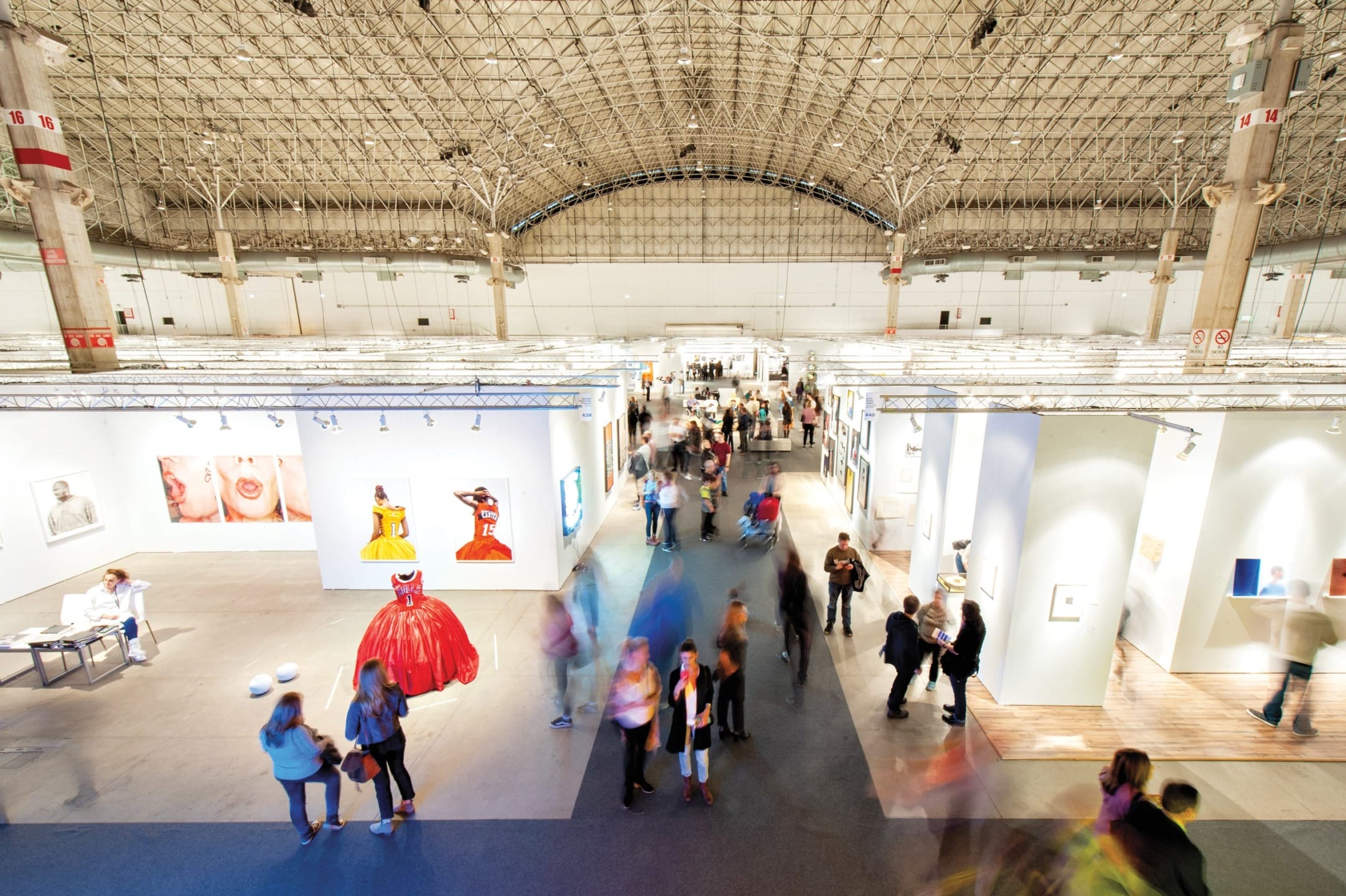 Art exhibition with visitors exploring various galleries and colorful artworks displayed in a large, well-lit hall.