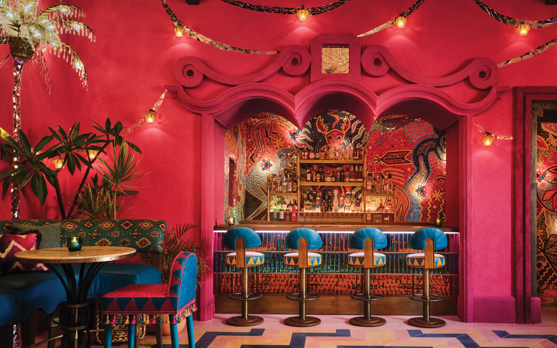 Vibrant bar interior with pink walls, eclectic decor, a colorful mural, and blue barstools lined up along the counter.