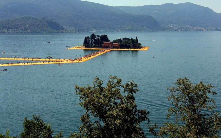 Floating walkway leading to island with trees on a lake, surrounded by mountains and clear blue sky in the background.