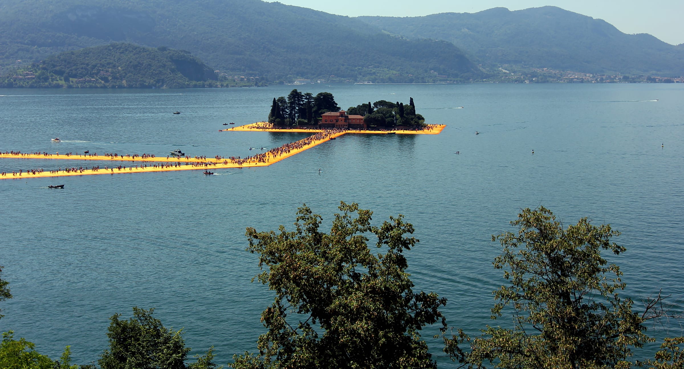 Floating walkways leading to a small island with trees on a lake surrounded by mountains.
