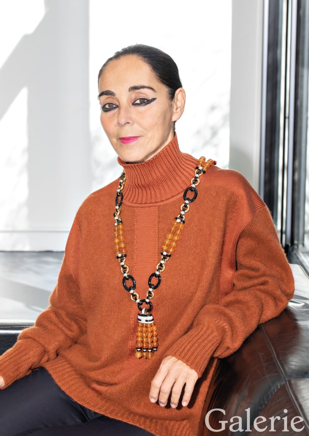 Person sitting in a bright room wearing an orange sweater and a long statement necklace, leaning against a couch.