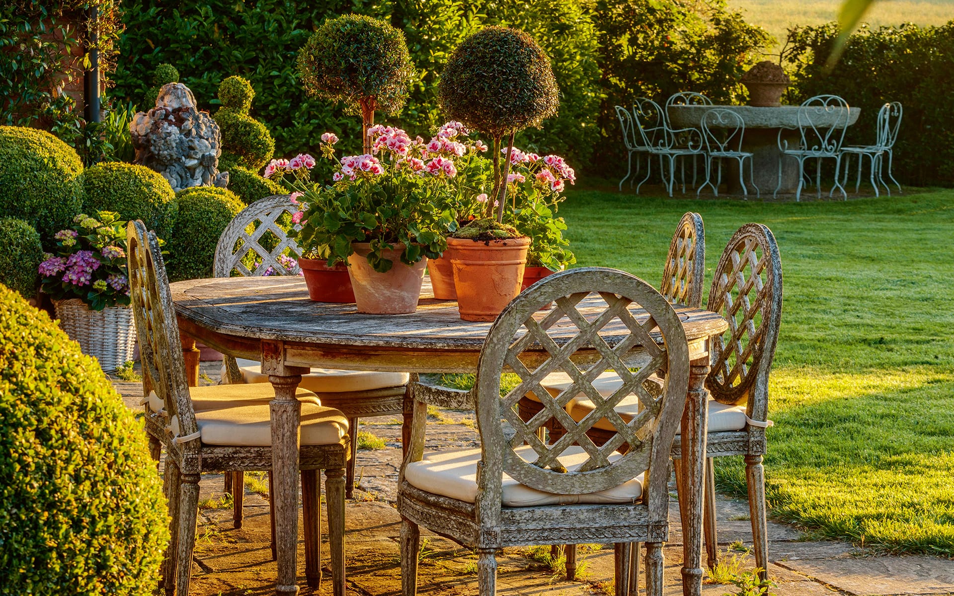 Rustic garden setting with a wooden table, potted flowers, and ornate chairs on a sunlit lawn.