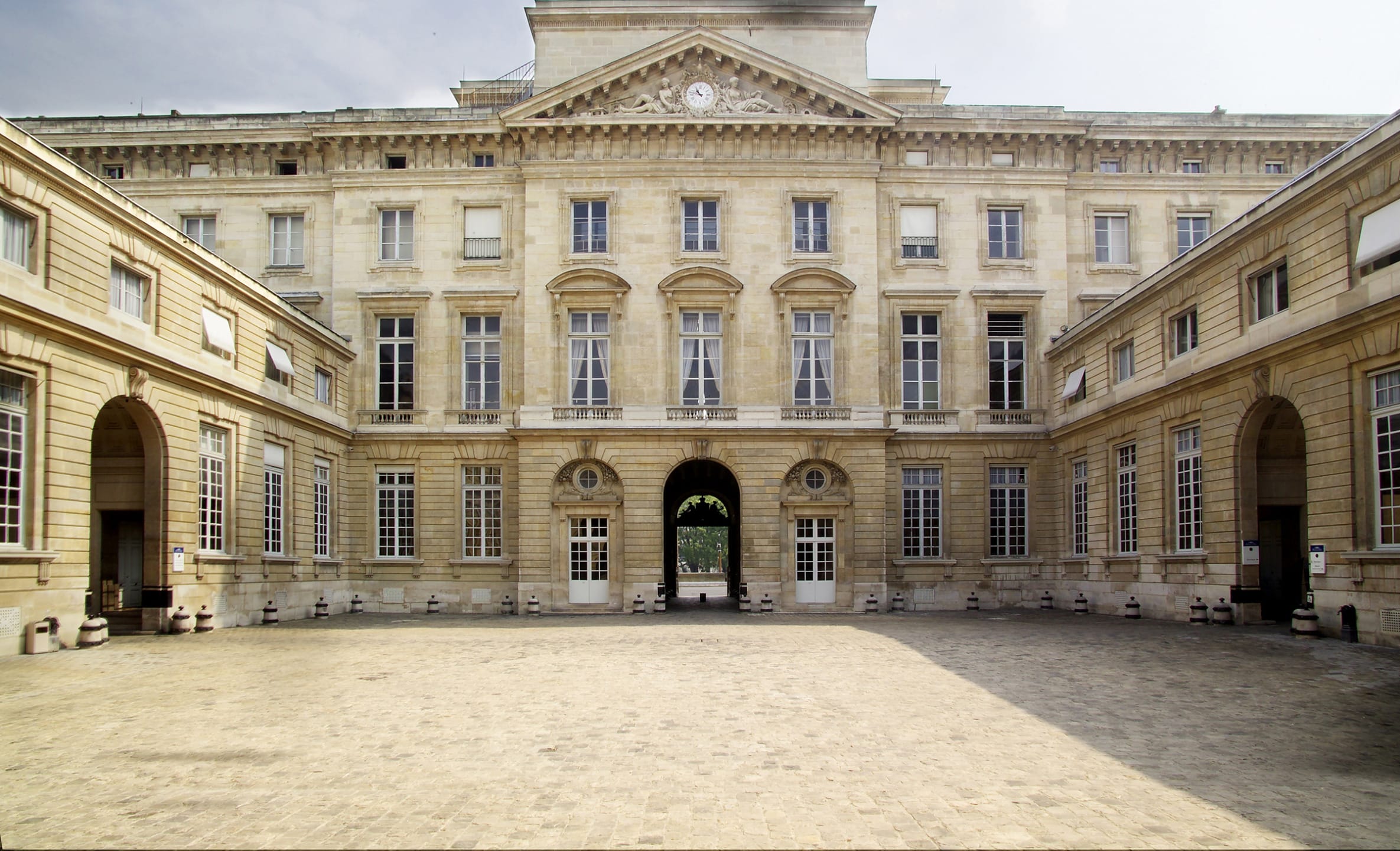 Historic stone building with a symmetrical facade and arched entrance, framed by two wings, under a partially cloudy sky.