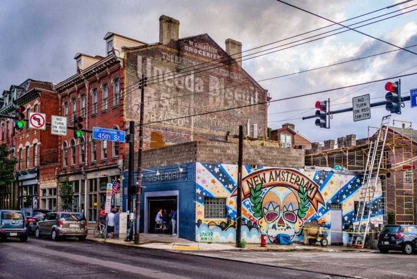 Street view of colorful graffiti on a building wall, with New Amsterdam text, cars, and street signs in an urban setting.