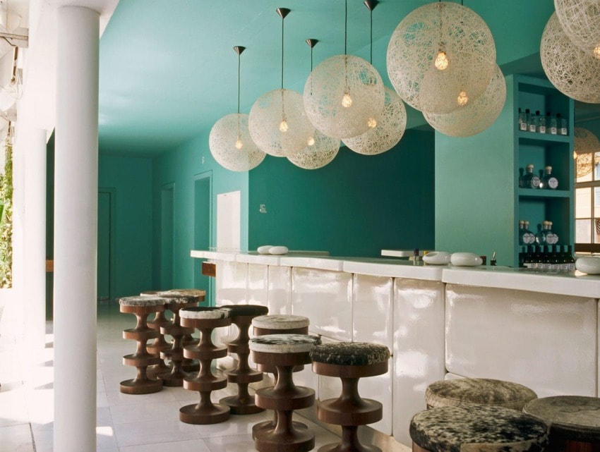 Modern bar with teal walls, unique light fixtures, wooden stools, and a sleek countertop.
