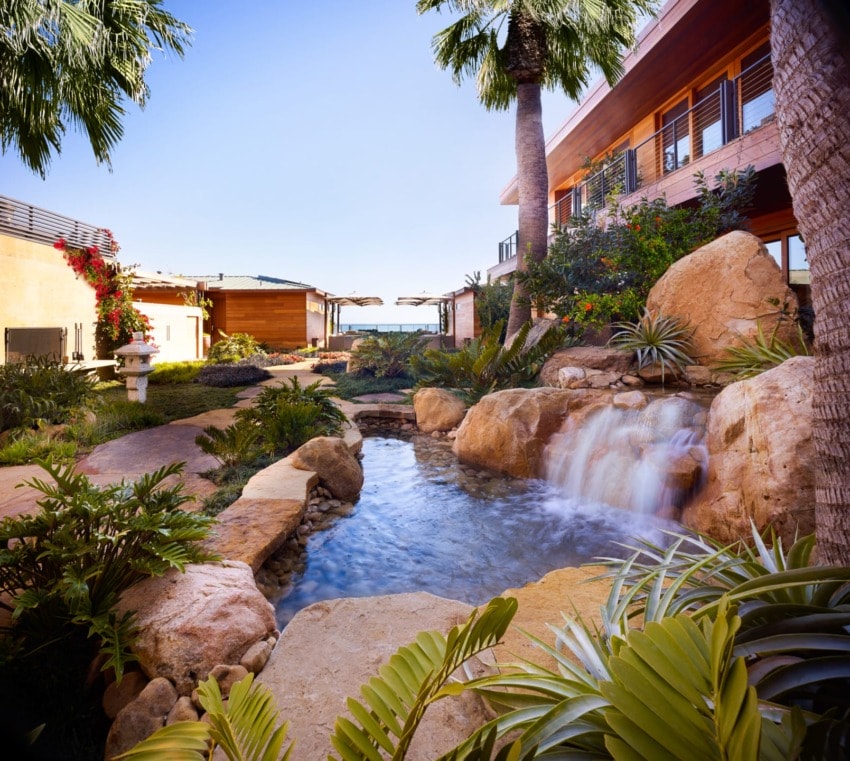 Tropical garden with a waterfall, surrounded by lush plants and modern buildings under a clear blue sky.