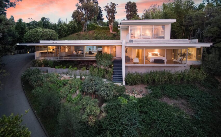 Modern hillside house with large windows surrounded by lush greenery at sunset.