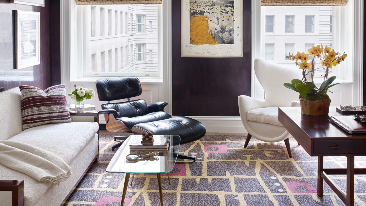 Modern living room with white sofa, black lounge chair, abstract rug, and large windows overlooking city buildings.