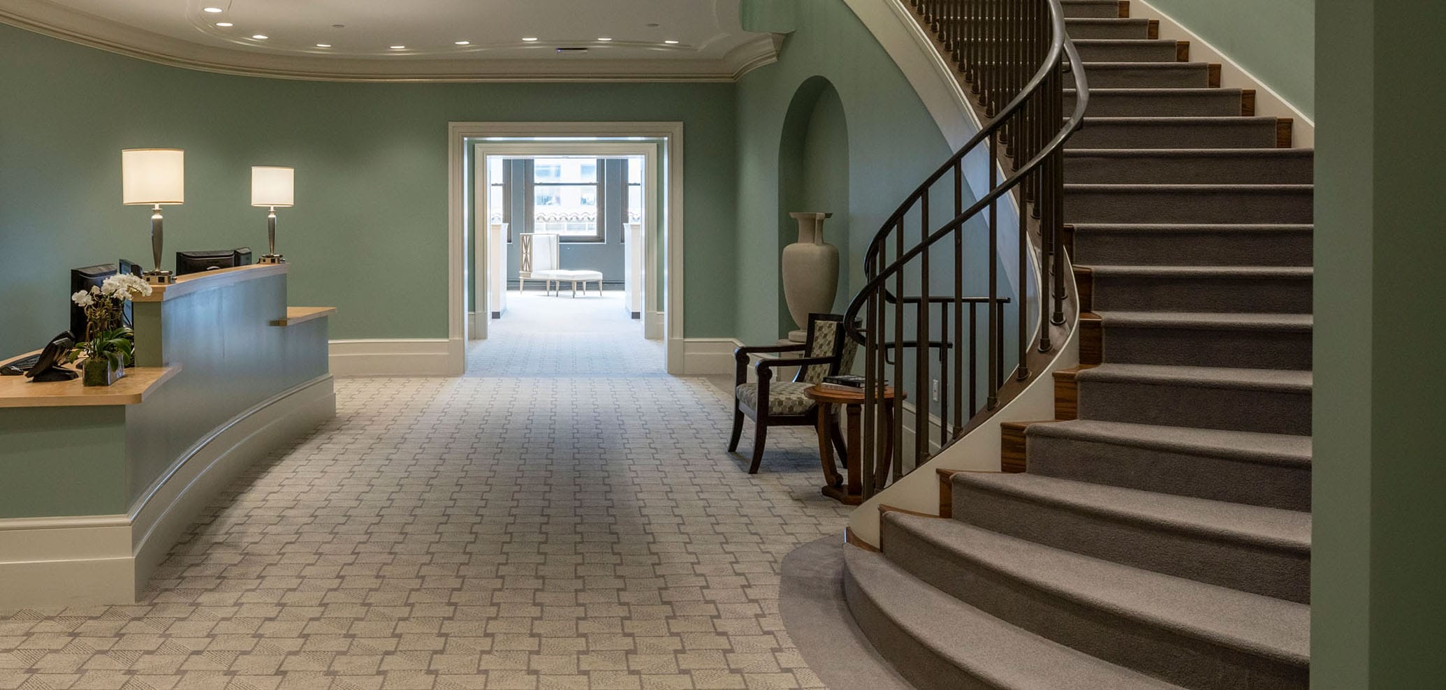 Hotel lobby with a curved staircase, decorative vase, and reception desk, featuring green walls and patterned carpet.