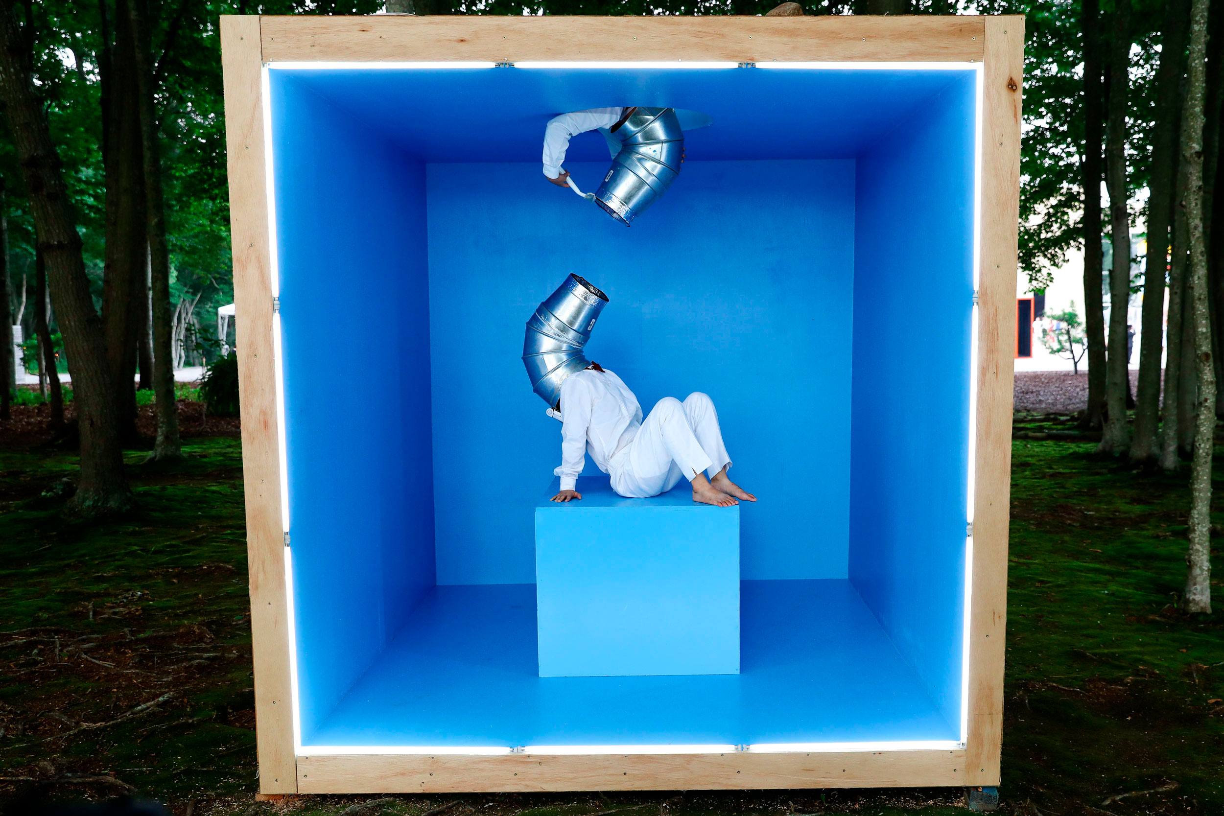 Person in white sitting inside a blue box with metallic tubes on head, surrounded by trees.