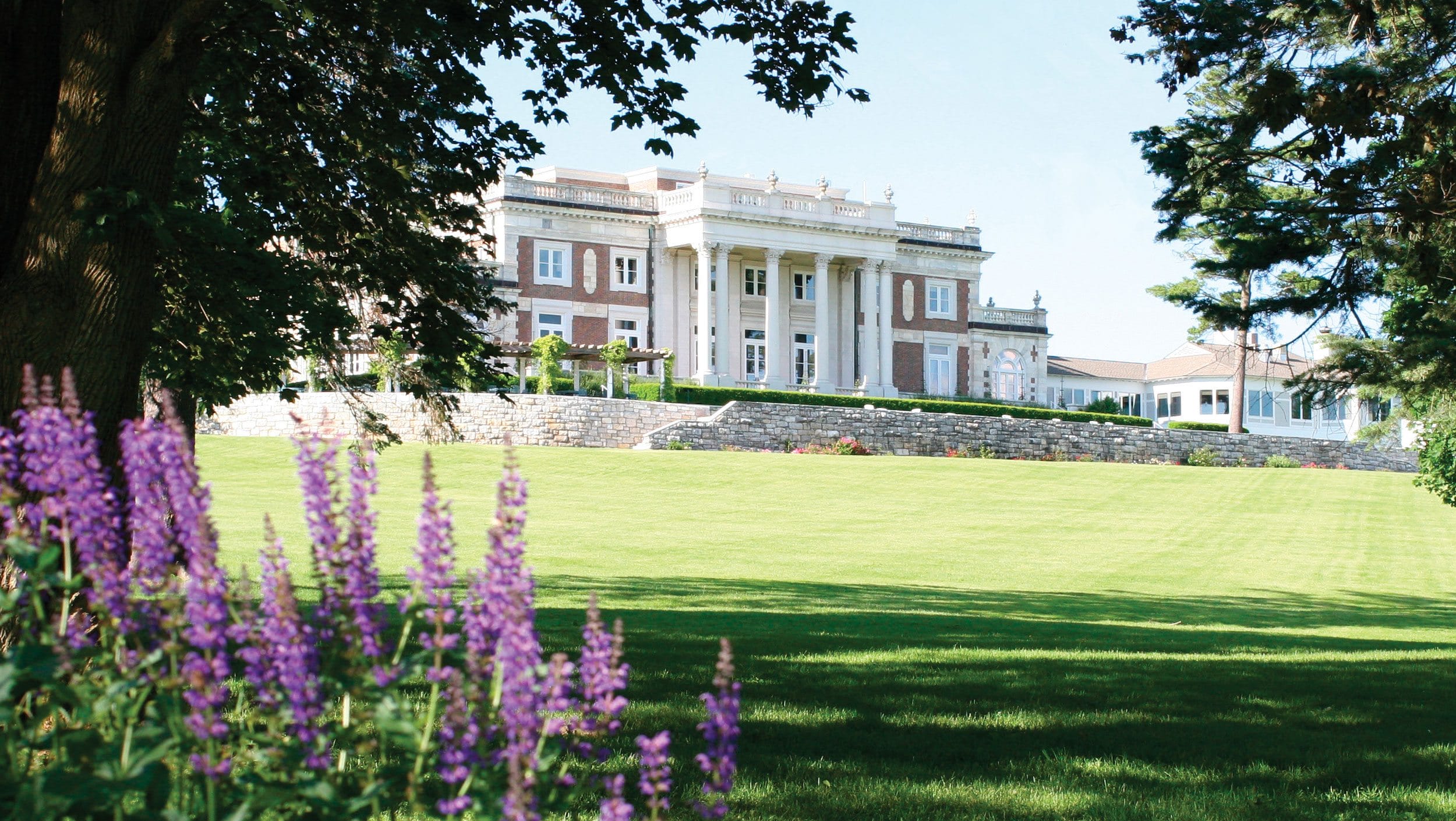 Historic mansion with grand columns surrounded by lush greenery and purple flowers in the foreground.
