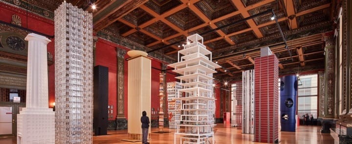 Museum exhibit featuring tall architectural models in an ornate hall with wooden floors and detailed ceiling.