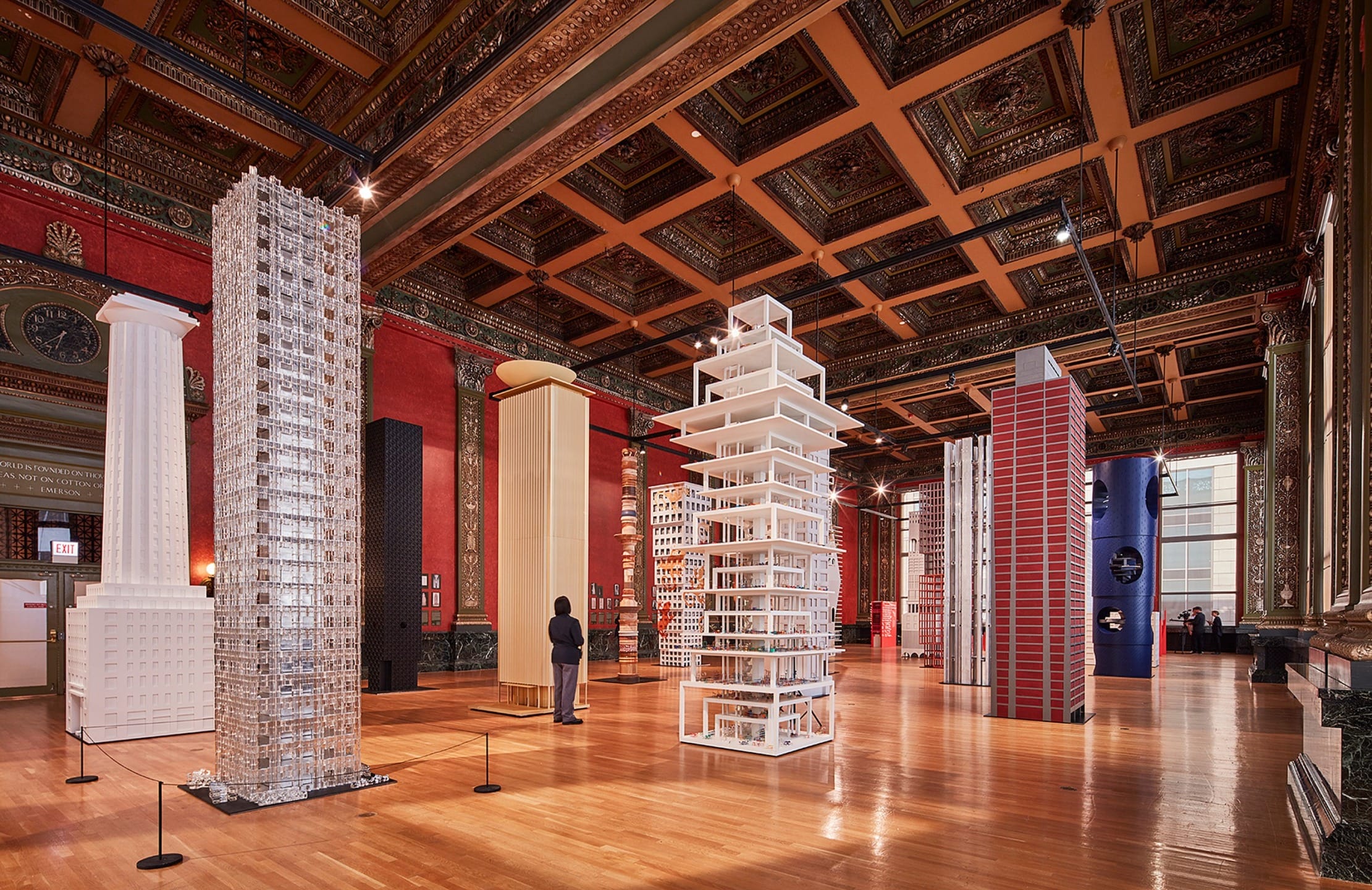 Museum exhibit featuring tall architectural models in an ornate hall with wooden floors and detailed ceiling.