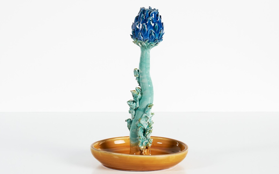 Unique ceramic sculpture of a tall, slender plant with a blue flower on top, displayed in a round, shallow, brown dish.
