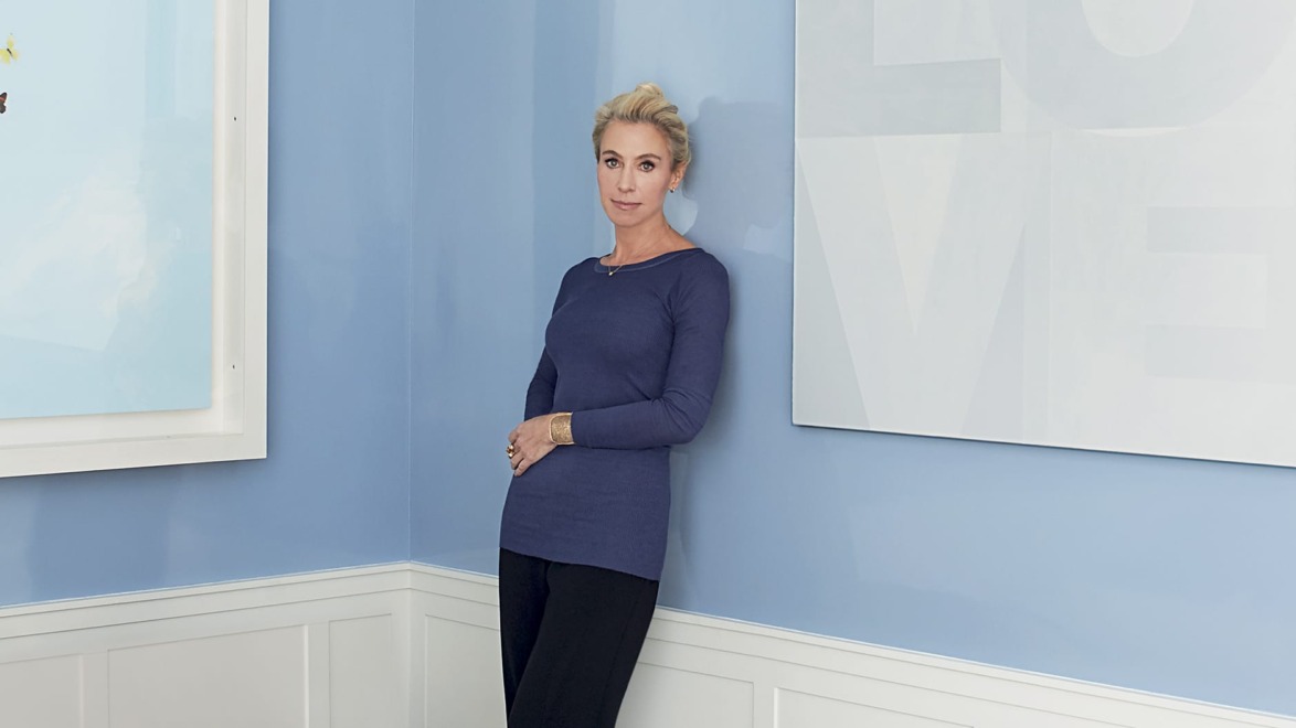 Person standing in a room with blue walls, wearing a blue top and black pants, next to two framed pieces of art.