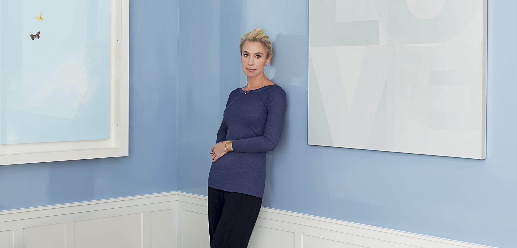 Person standing in a room with blue walls, wearing a blue top and black pants, next to two framed pieces of art.