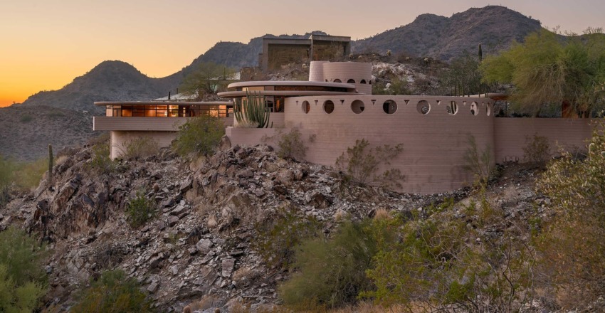 Desert house with circular design elements on rocky hillside with mountains in the background at sunset.