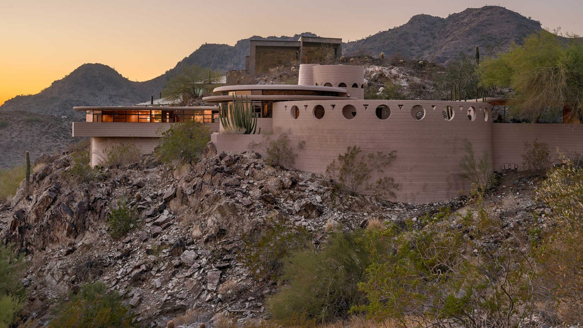 Desert house with circular design elements on rocky hillside with mountains in the background at sunset.