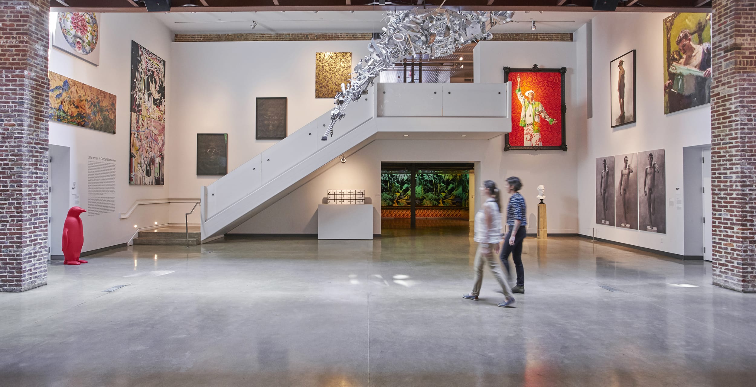 Modern art gallery interior with various artworks on display, featuring a staircase and two people walking across the polished floor.