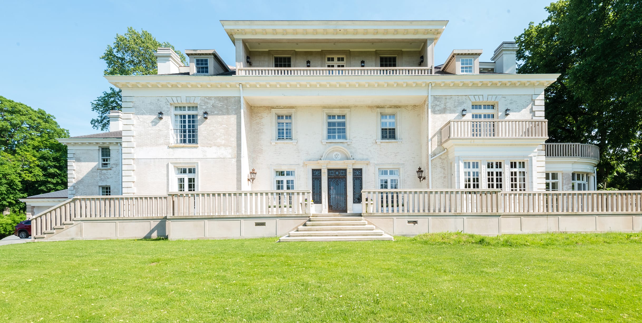 Large white mansion with columns, multiple balconies, and a well-maintained lawn under a clear blue sky.