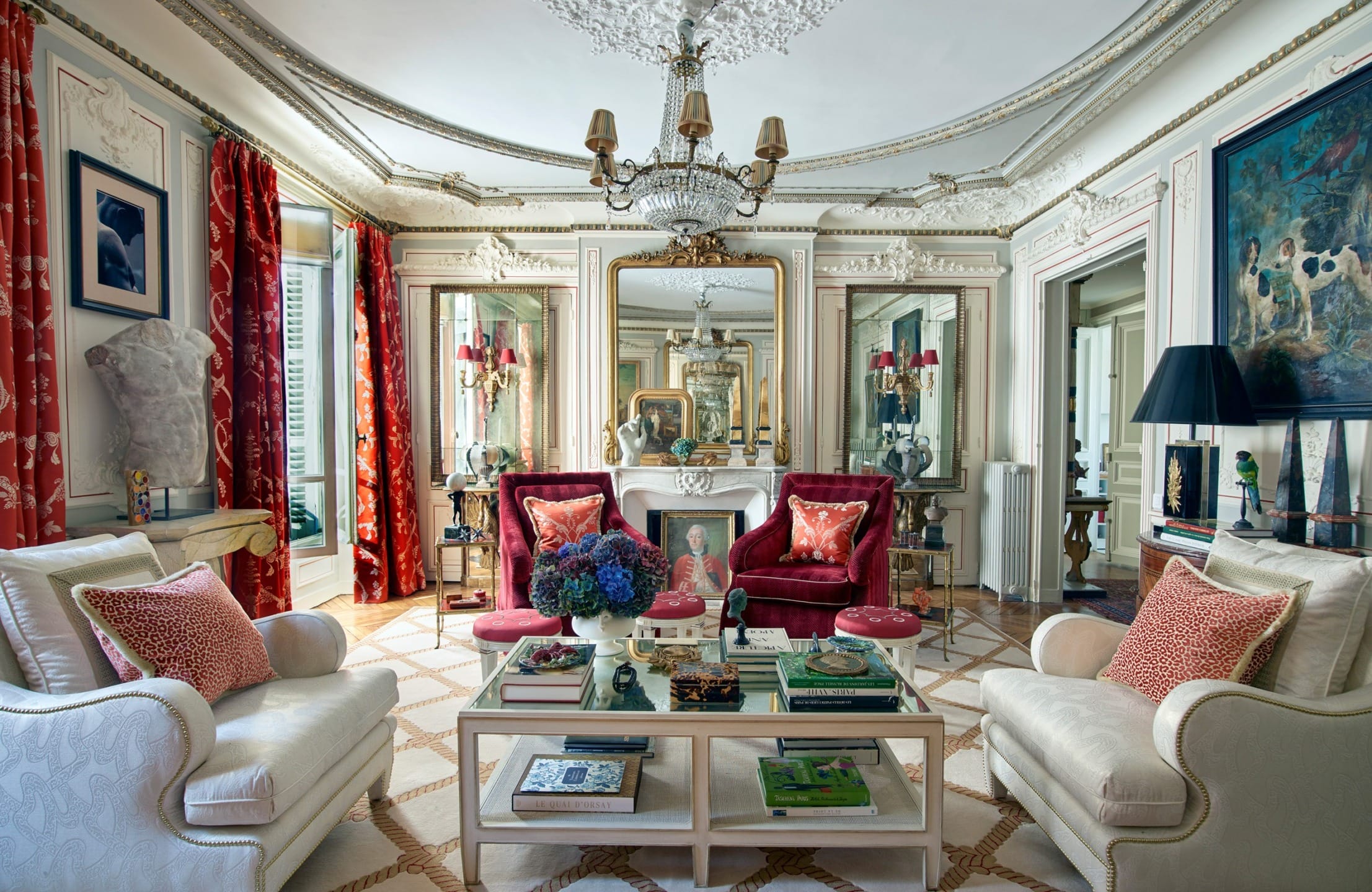 Elegant living room with vintage decor, chandeliers, red velvet furniture, and patterned floor, framed by rich red curtains.