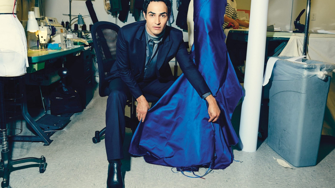 Person in a tailor shop, kneeling beside a vibrant blue dress on a mannequin, with sewing materials in the background.