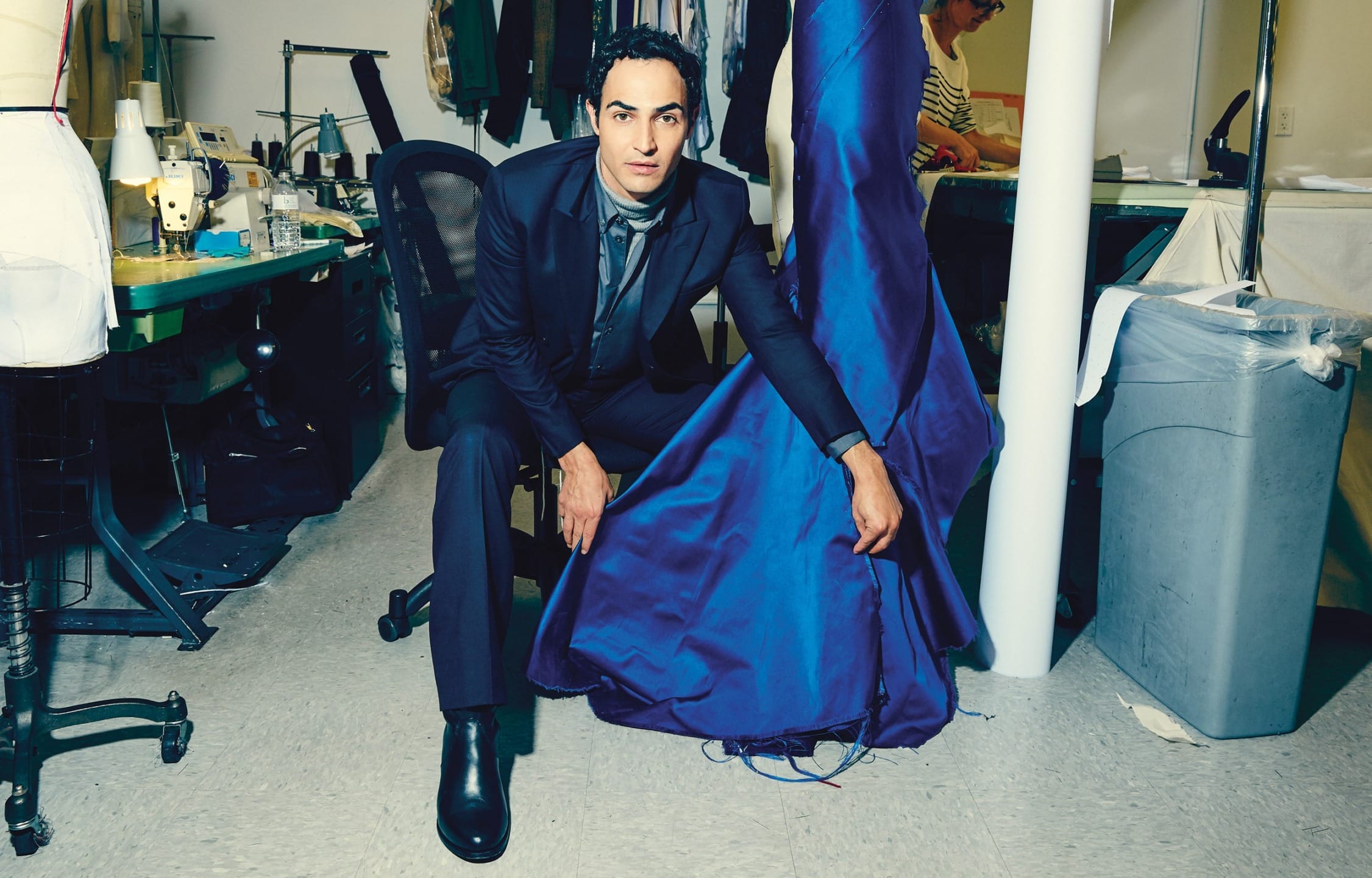 Person in a tailor shop, kneeling beside a vibrant blue dress on a mannequin, with sewing materials in the background.