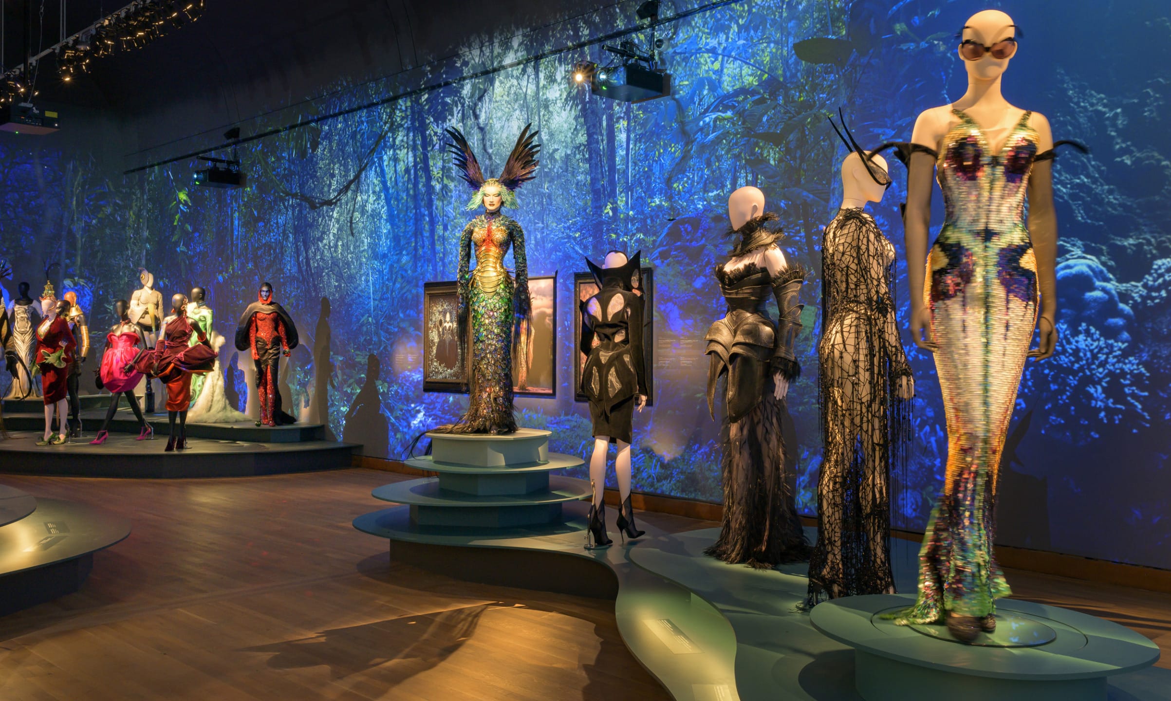 Exhibition of colorful, avant-garde fashion designs on mannequins in a dimly lit room with a blue aquatic-themed backdrop.