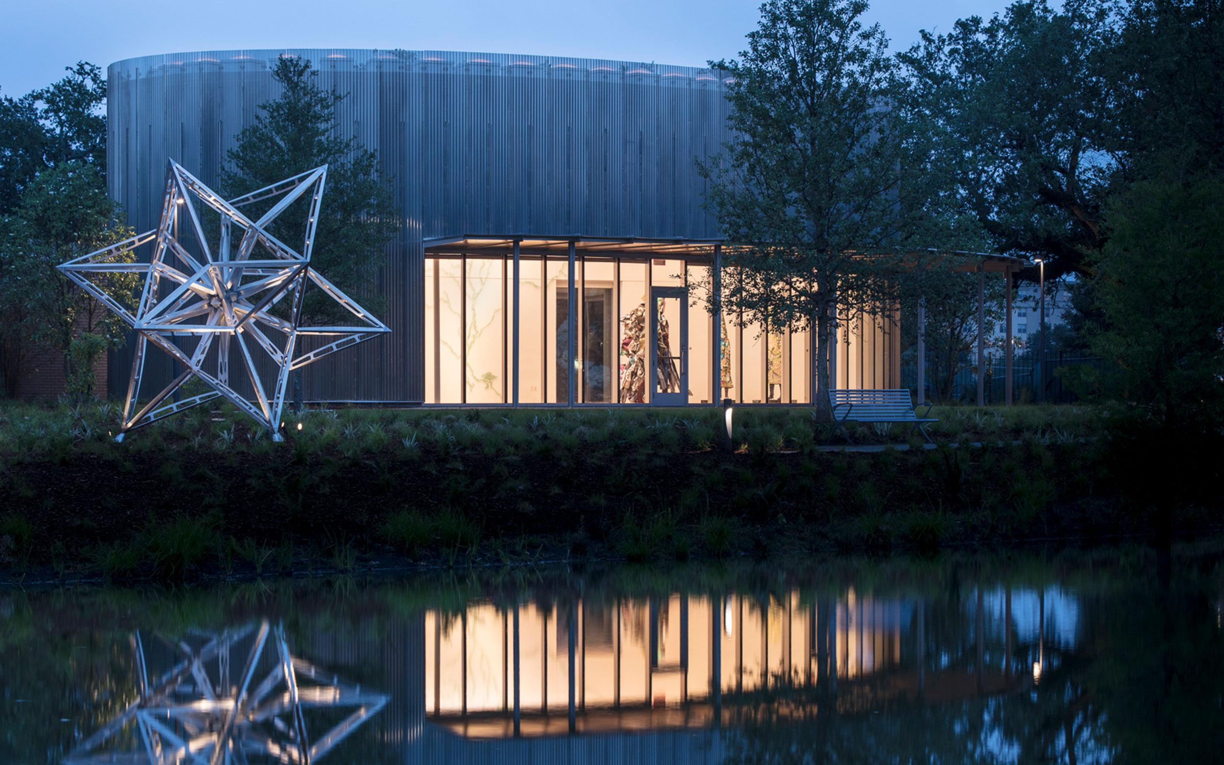 Contemporary building with large geometric star sculpture and glass facade reflected in a tranquil pond at dusk.