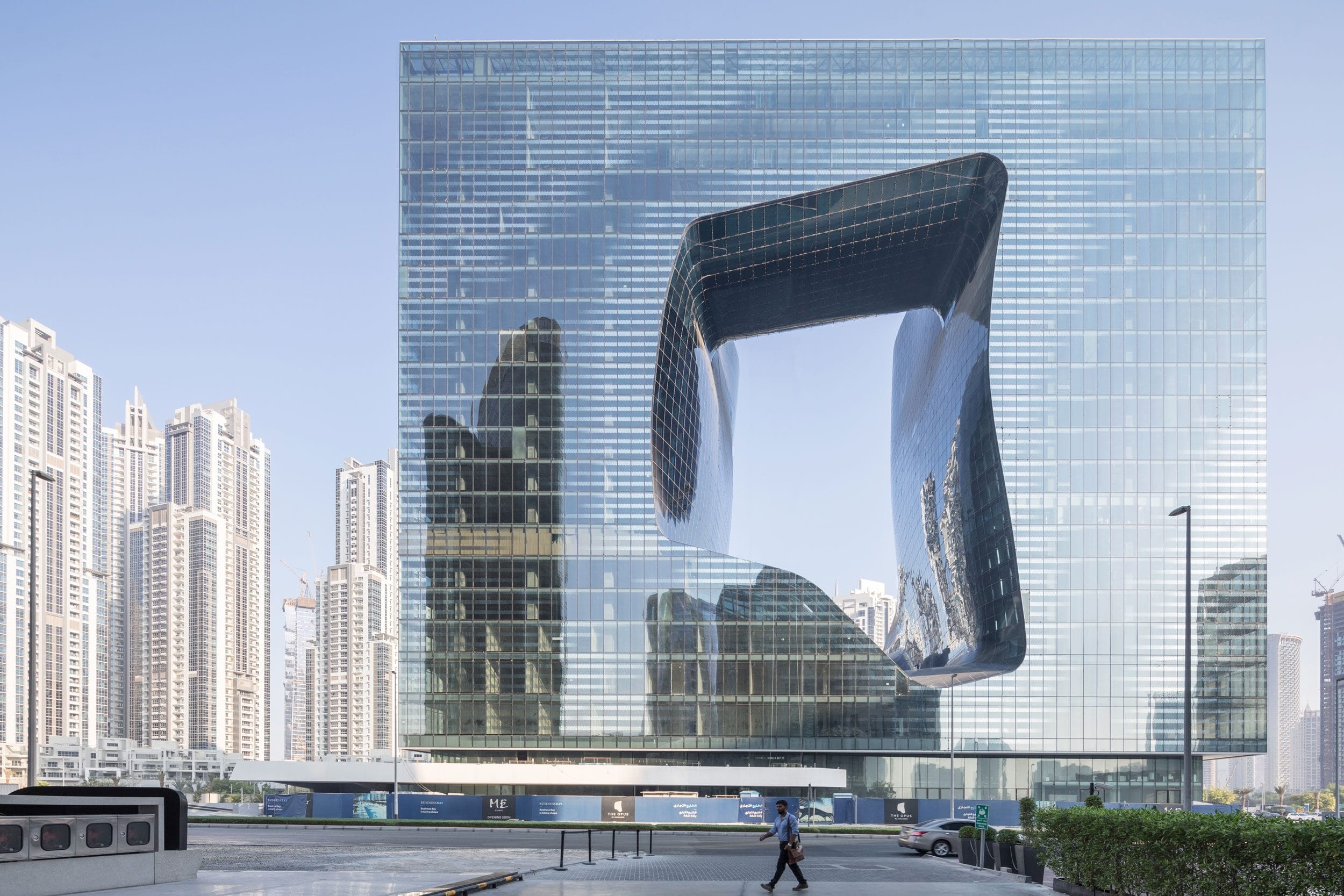 Modern glass building with unique architectural cutout, surrounded by cityscape and skyscrapers on a clear day.