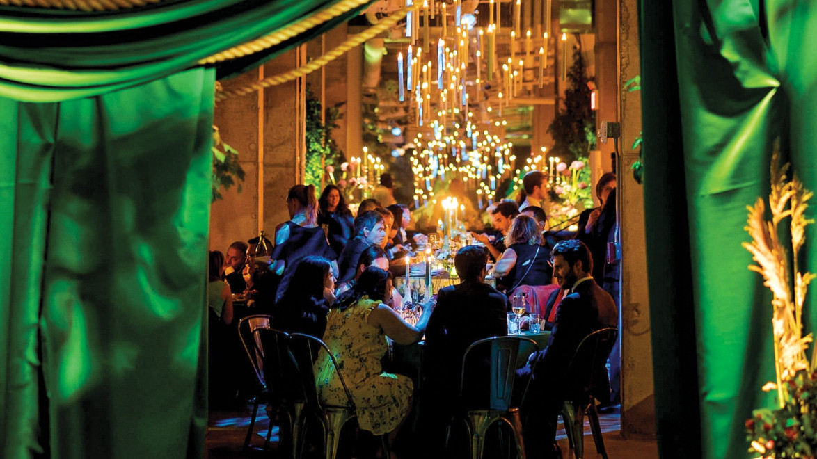 Guests dining in a warmly lit room with green drapery and glowing chandeliers overhead.