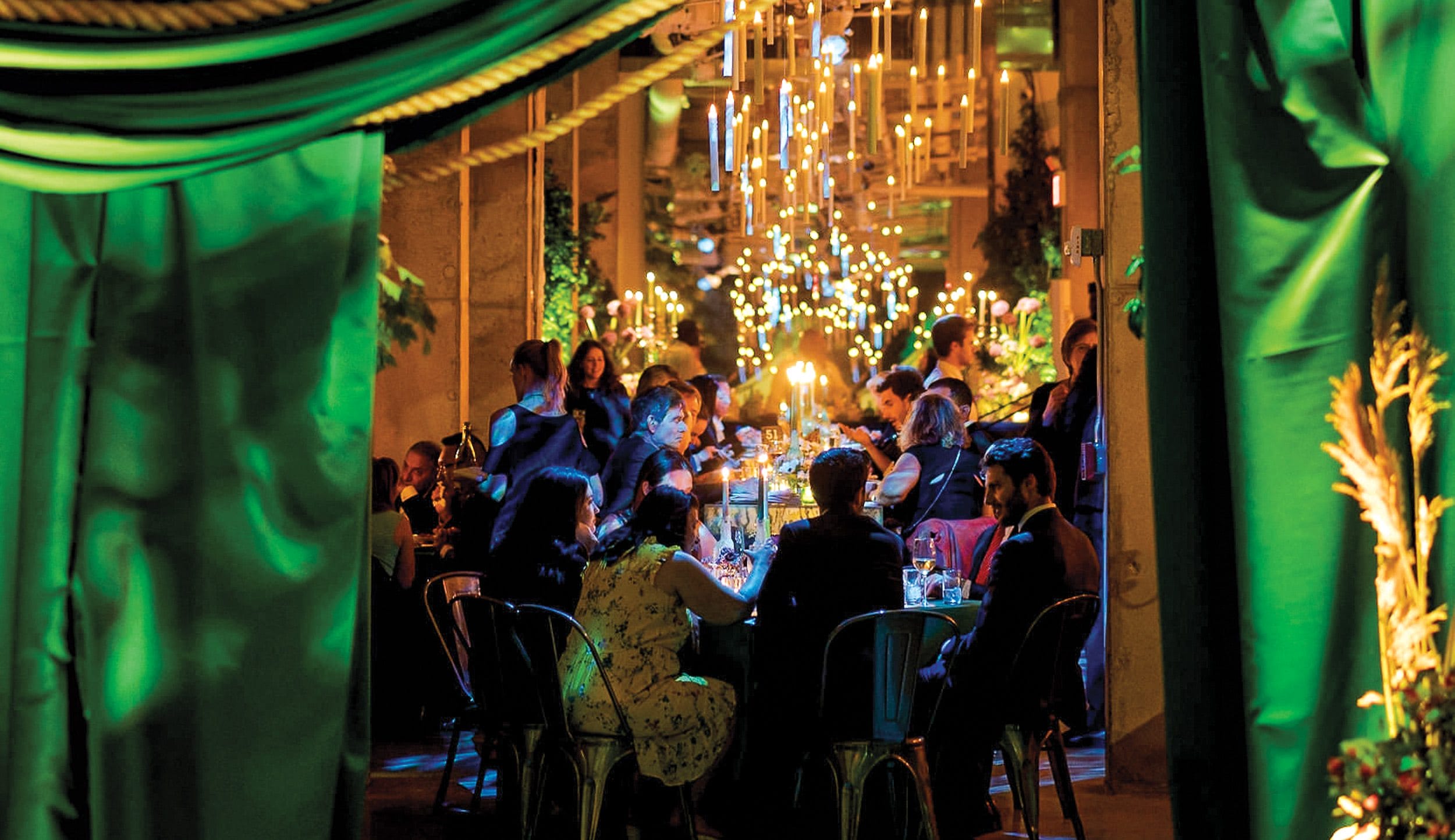 Guests dining in a warmly lit room with green drapery and glowing chandeliers overhead.