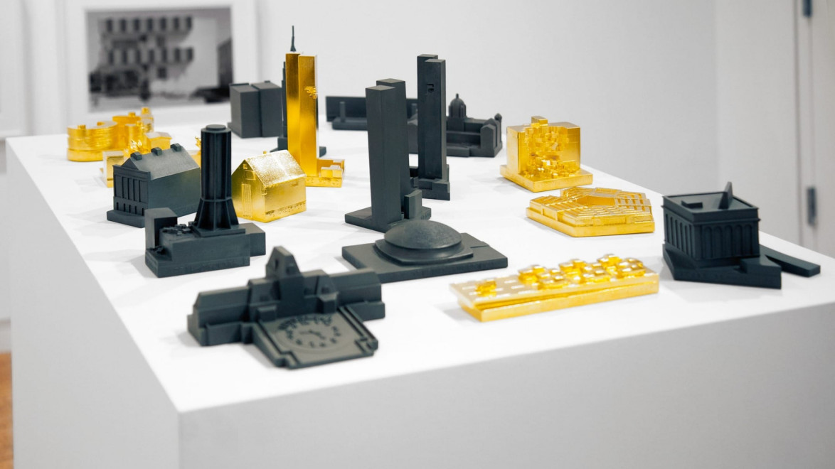 Miniature cityscape models in black and gold displayed on a white pedestal in an art gallery setting.