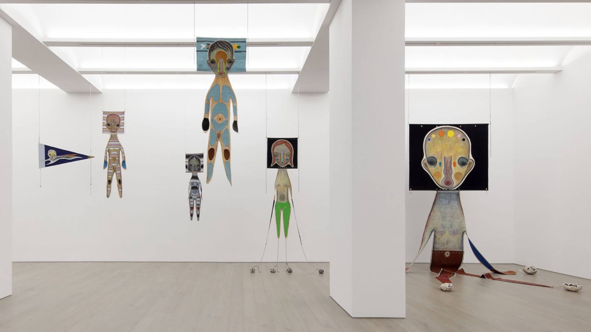 Art installation with hanging abstract humanoid figures in a modern gallery with white walls and wooden floor.