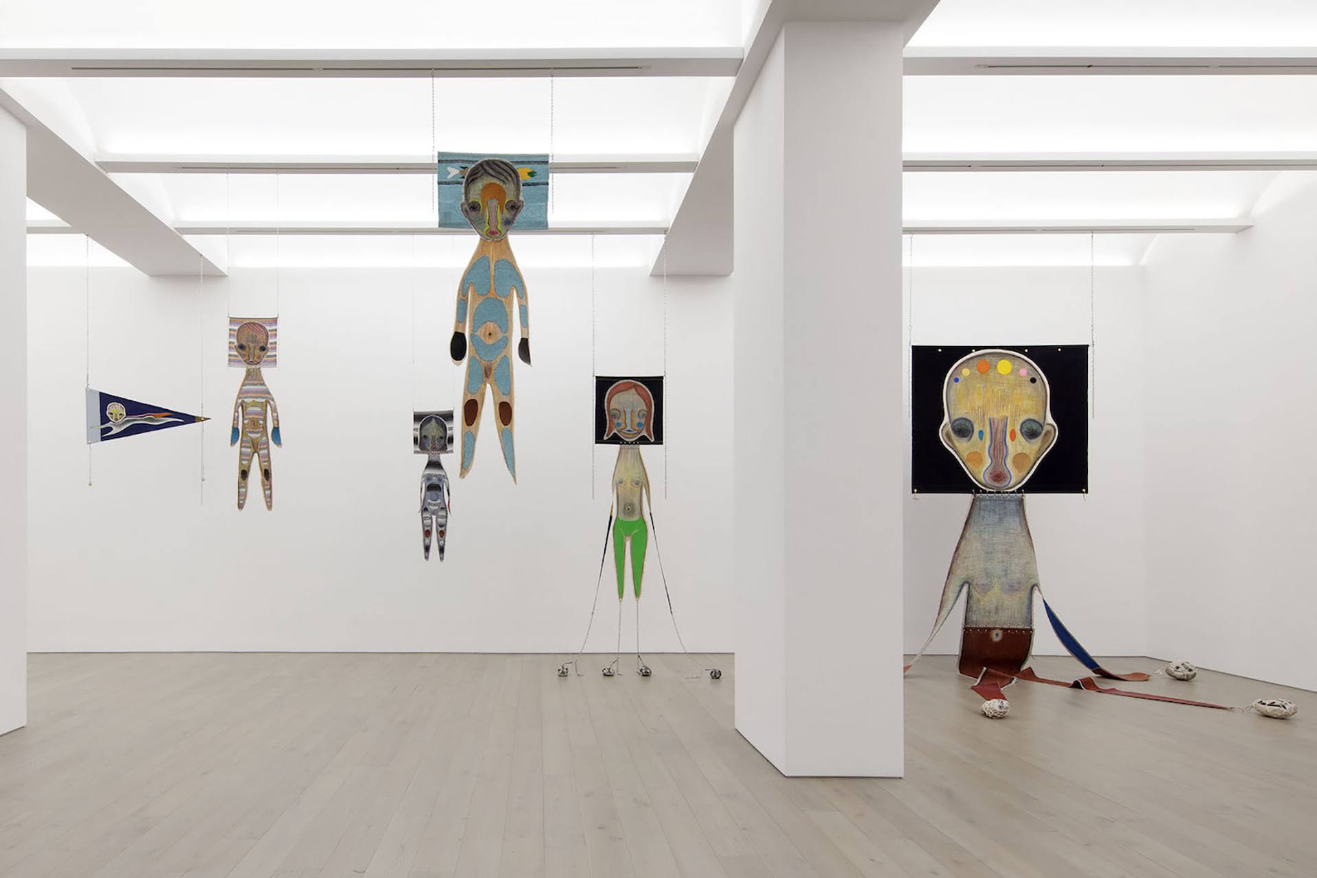 Art installation with hanging abstract humanoid figures in a modern gallery with white walls and wooden floor.