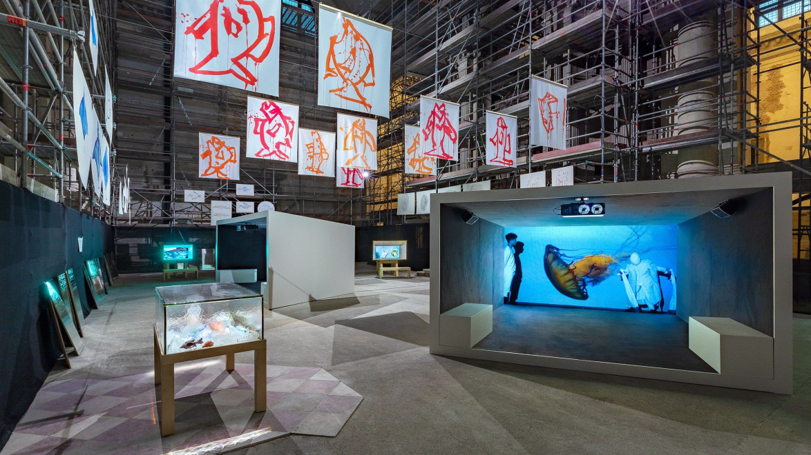 Art exhibit with digital screens, illuminated displays, and hanging banners in an industrial space with scaffolding.