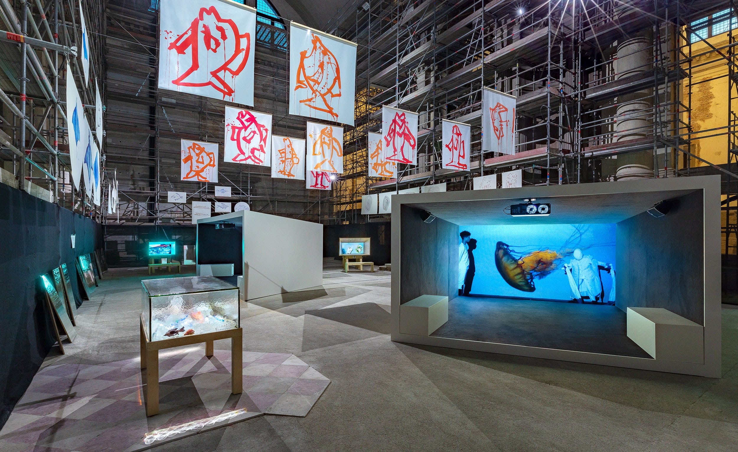 Art exhibit with digital screens, illuminated displays, and hanging banners in an industrial space with scaffolding.