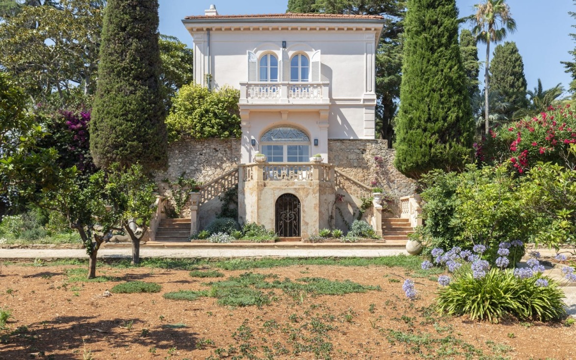 Elegant two-story villa surrounded by trees and plants with arched windows and a stone pathway in front.