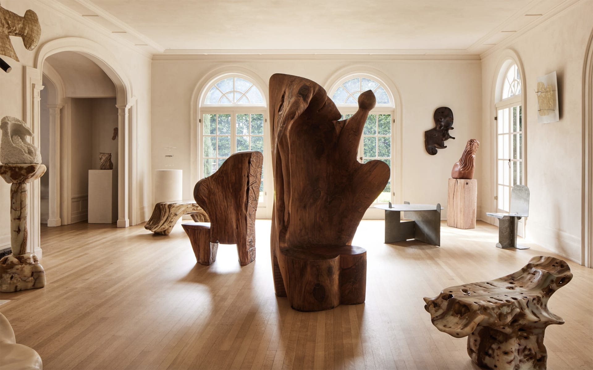Gallery with wooden sculptures and marble artworks displayed in a spacious, naturally lit room with large arched windows.