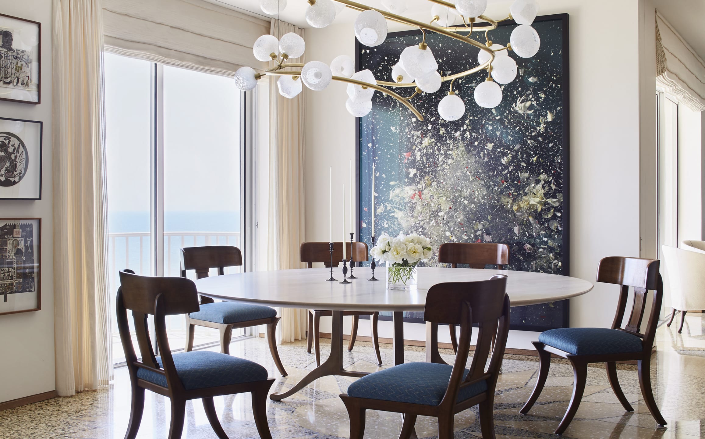 Dining room with a round table, blue cushioned chairs, modern chandelier, and a large abstract painting on the wall.