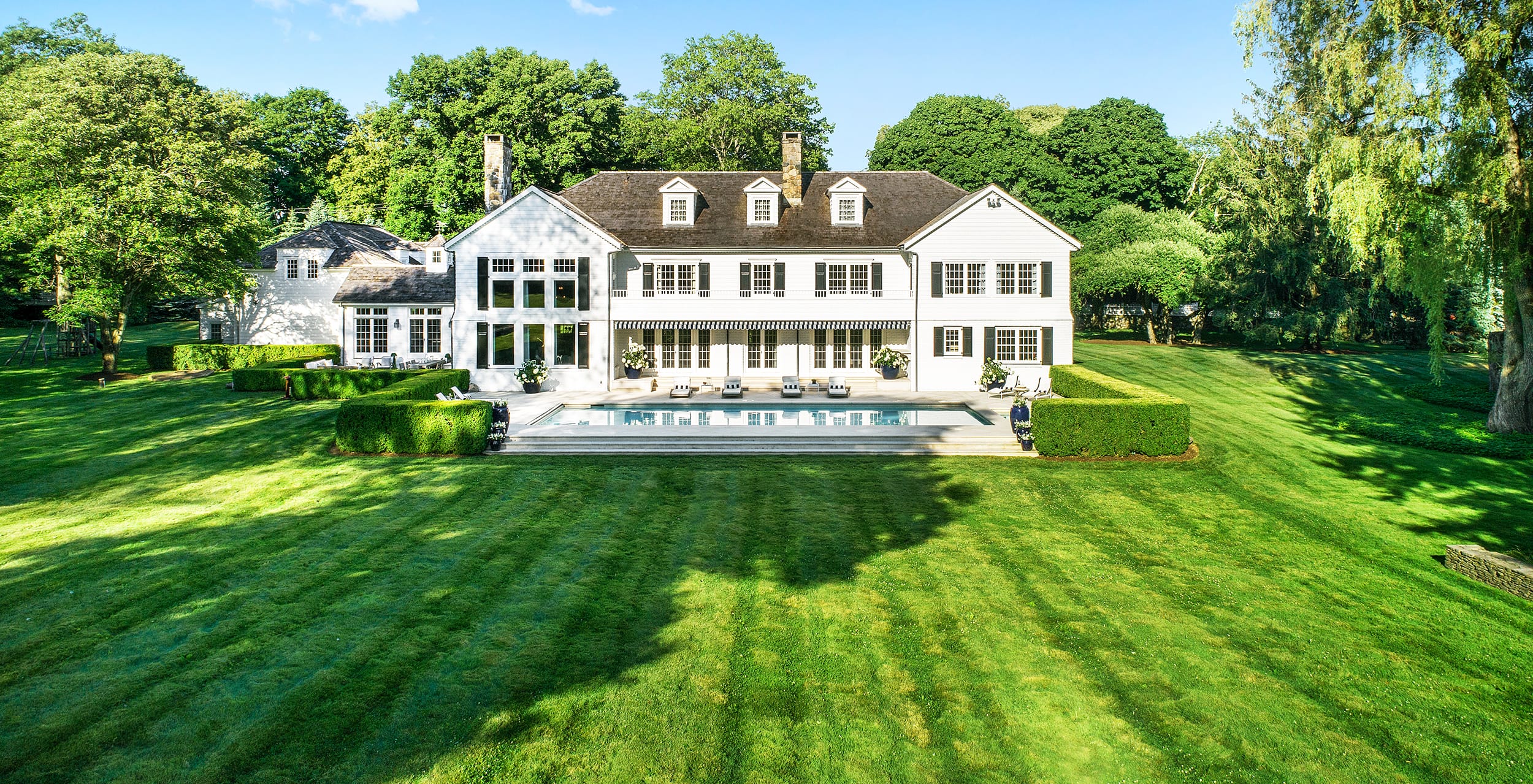 Spacious white house with large windows, set in a lush green lawn, featuring a backyard pool and surrounded by trees.