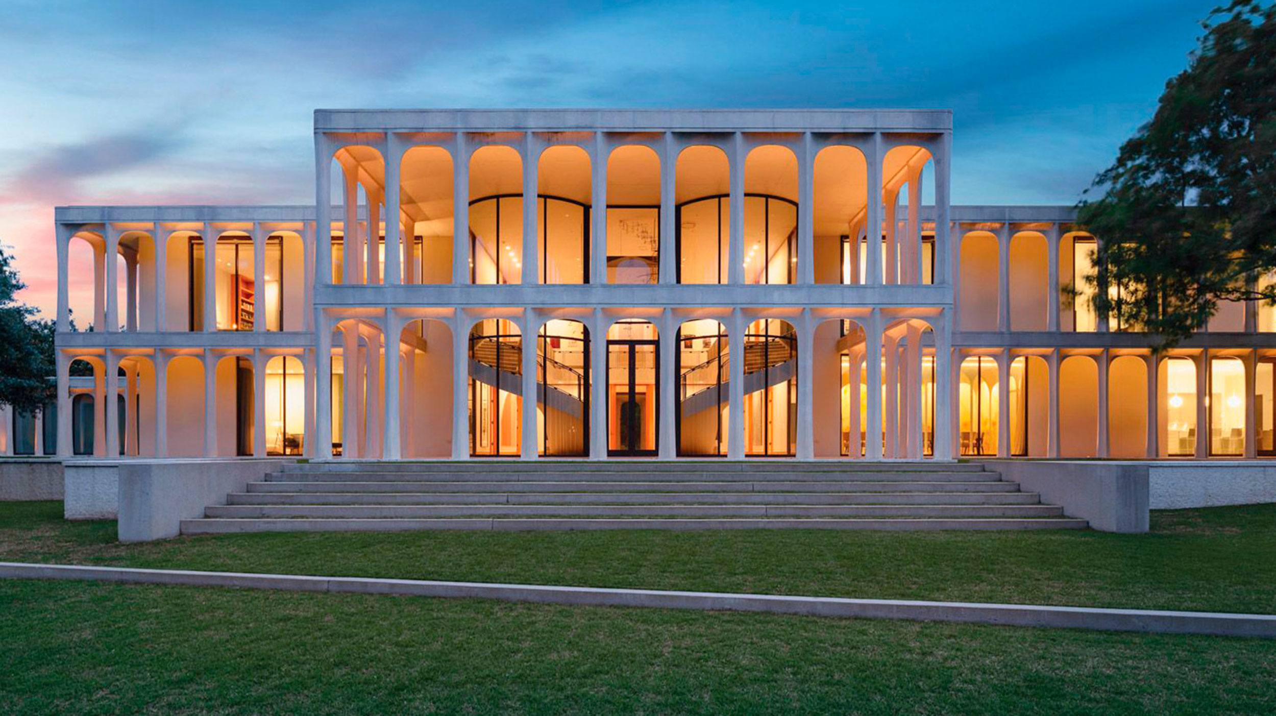 Modern building with illuminated windows, featuring multiple arches and columns, set against a twilight sky and surrounded by grass.